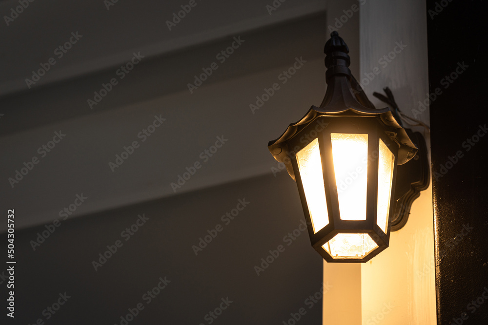 A classic style street lighting lamp is glowing in orange warm light shade in evening night time with background of building structure. Photo contained noise due to dark condition. Selective focus.
