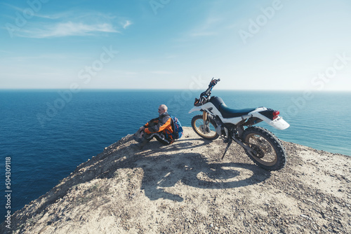 Elderly man resting in enduro motorcycle trip on mountain cliff with amazing sea skyline view