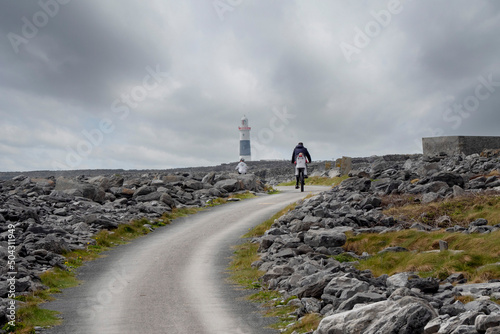 Family with children cycling on small narrow country road in rough stone terrain to a lighthouse. cloudy sky. Inisheer  Aran island  county Galway  Ireland. Irish landscape. Adventure concept.