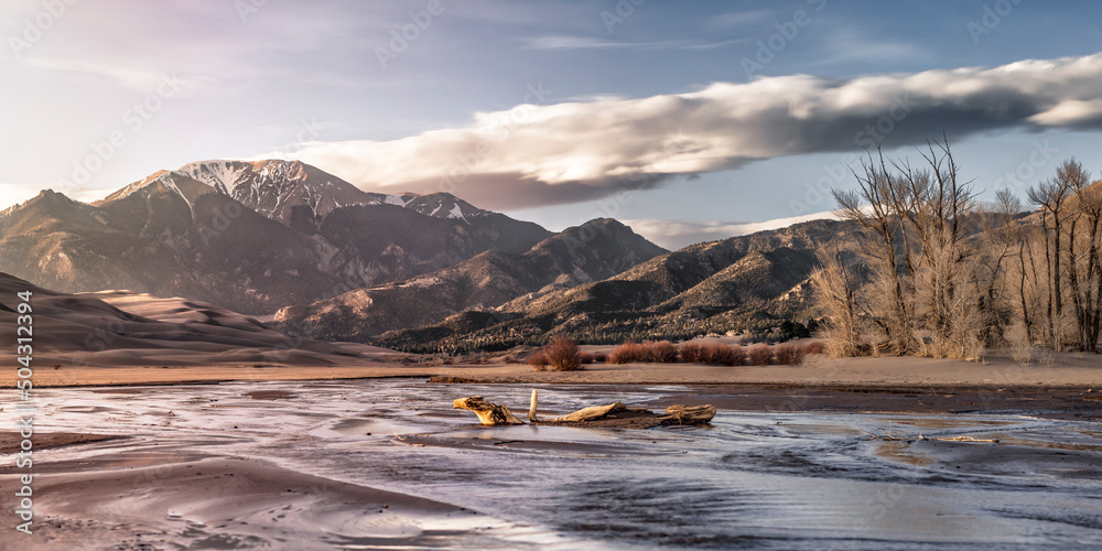 A Colorado landscape scene of sand dunes, a river, and snow capped mountains in the distance. 