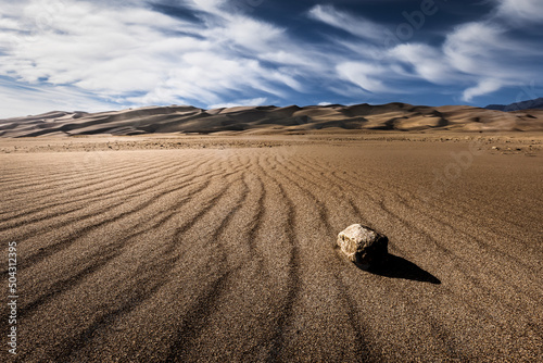Fototapeta Naklejka Na Ścianę i Meble -  A rock in the sand with sand dunes in the distance. There are wispy clouds visible in the sky. The landscape scene is very brown and dry.