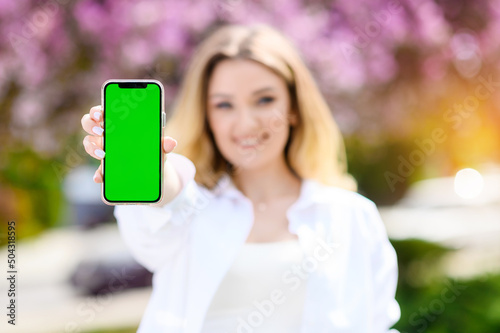 Beautiful blonde hair girl in a white shirt holding a phone with a green screen on a background of the park