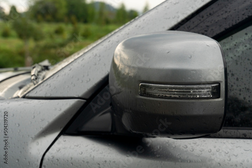 Close up side mirror wing cover material of gray car is foldable parking time. by automatic from system. on surface car wet water are raindrops from outdoor parking while it rains.