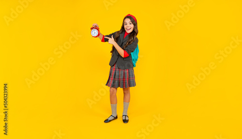 amazed teen girl with school bag pointing finger on alarm clock on yellow background, time. © be free
