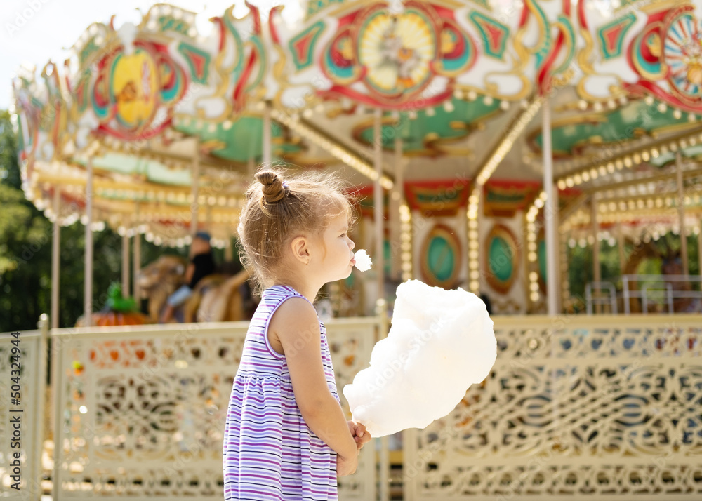 Fototapeta premium Funny blonde girl in colorful dress eating cotton candy at the carnival