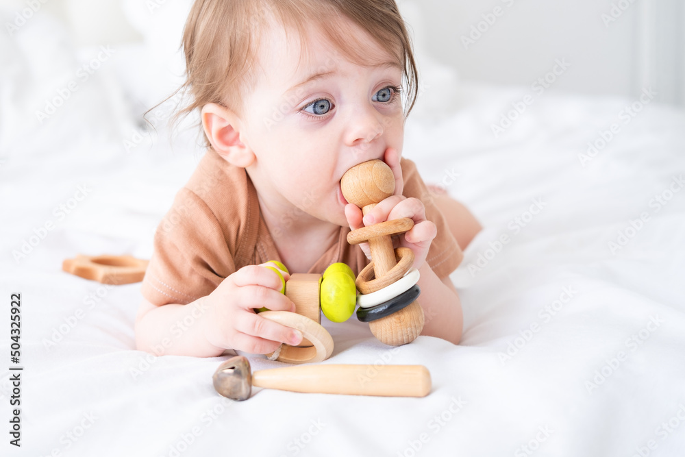 baby girl with blue eyes in brown bodysuit playing with wooden toys on white bedding on bed baby girl with blue eyes in brown bodysuit playing with wooden toys on white bedding on bed