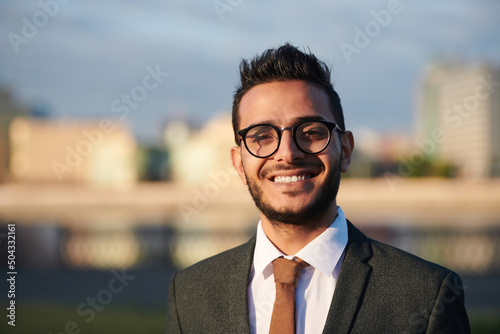 Portrait of cheerful successful young middle-eastern entrepreneur in eyeglasses standing against cityscape
