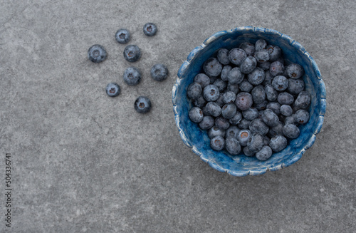 On top shot of a homemade blue clay bowl filled with fresh blueberries. There are also a few berries on the gray stone background. There is space for text on the side.
