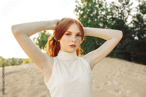 Unemotional woman with freckles touching long red hair and looking at camera while standing on sandy hill in summer evening  photo