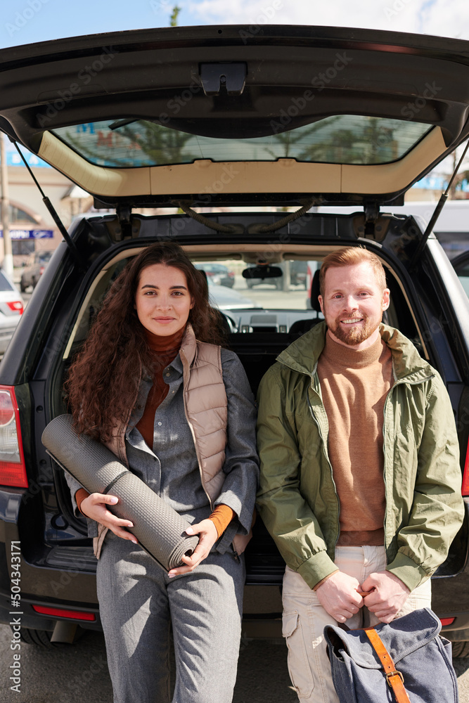 Portrait of content beautiful young couple holding satchel and mat while leaning on car trunk outdoors