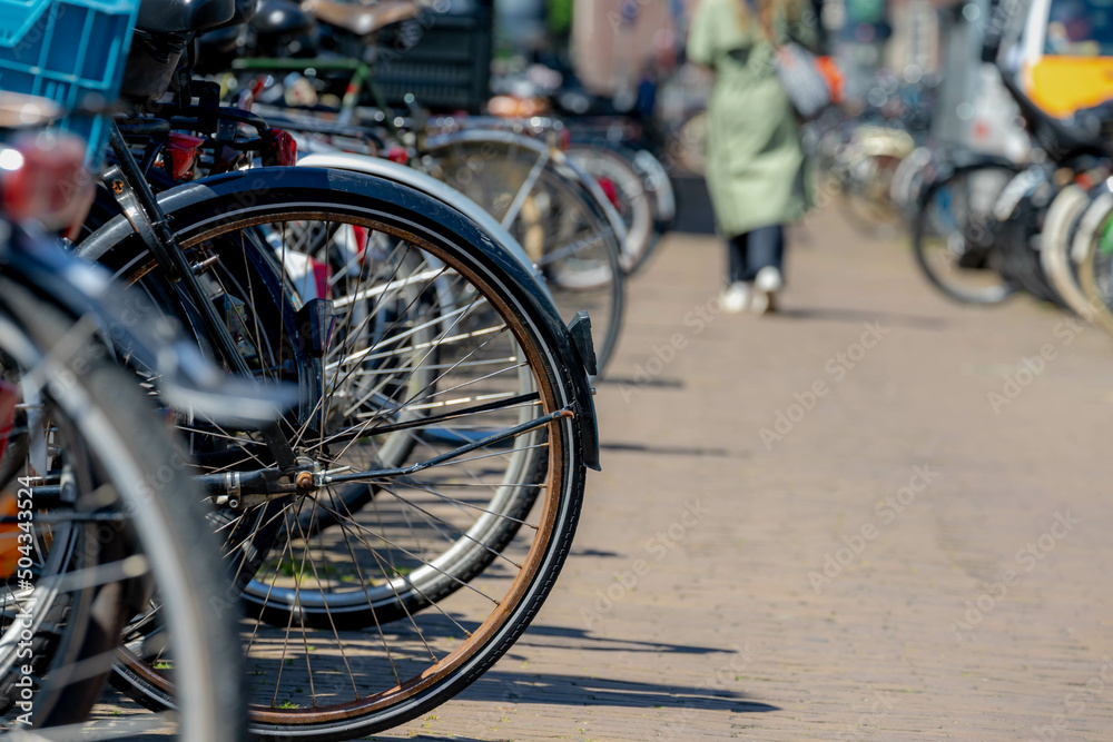 Selective focus of back wheel of bicycle in the row, Outdoor public parking area with blurred people walking in parking slot, Cycling is a common mode of transport in the Netherlands. Selective focus of back wheel of bicycle in the row, Outdoor public parking area with blurred people walking in parking slot, Cycling is a common mode of transport in the Netherlands.