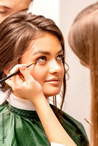The makeup artist applies a concealer under the eyes using a makeup brush