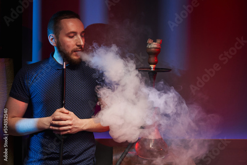 a young man smokes a hookah in an atmospheric hookah bar illuminated by color filters and exhales a cloud of white smoke