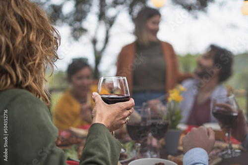 Blonde woman holding a glass of red wine talking with friends at picnic table in the countryside - focus on the hand, defocused unrecognizable people in the background - wine tasting lifestyle concept