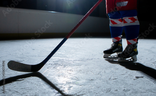 Man hockey player in sports uniform and skates standing on ice arena with stick in his hands. An athlete hones his skills in playing hockey on ice of stadium in dark with backlight. Legs close up.