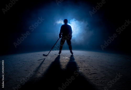 Fototapete Dark silhouette of a male hockey player in a uniform, helmet and skates with a stick on the ice arena with smoke and blue back light