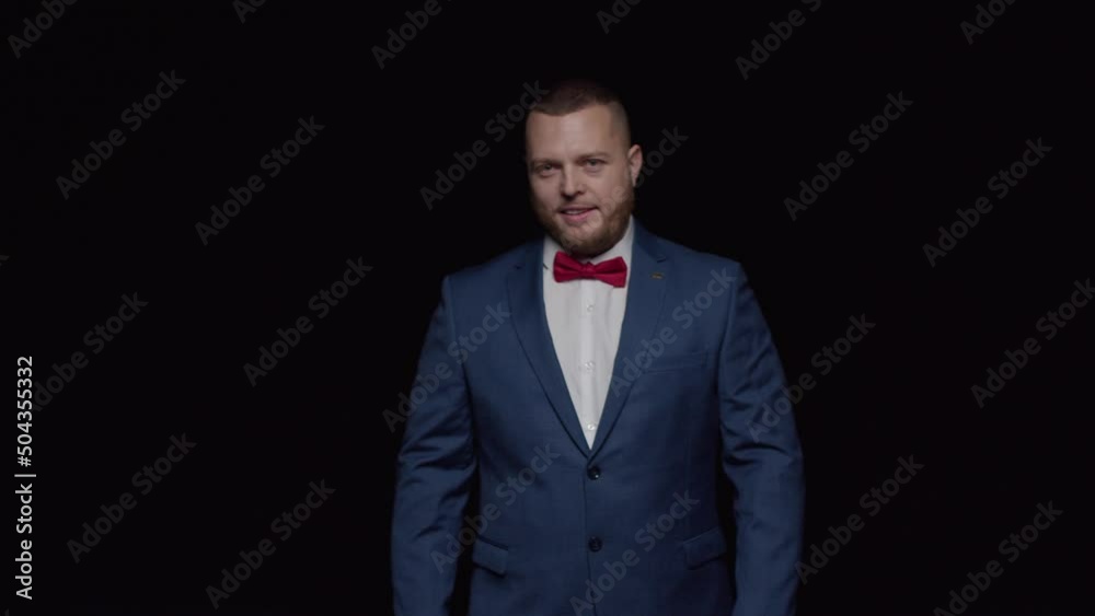 An elegant man in a blue tuxedo and a red tie smiles sweetly against a black background.