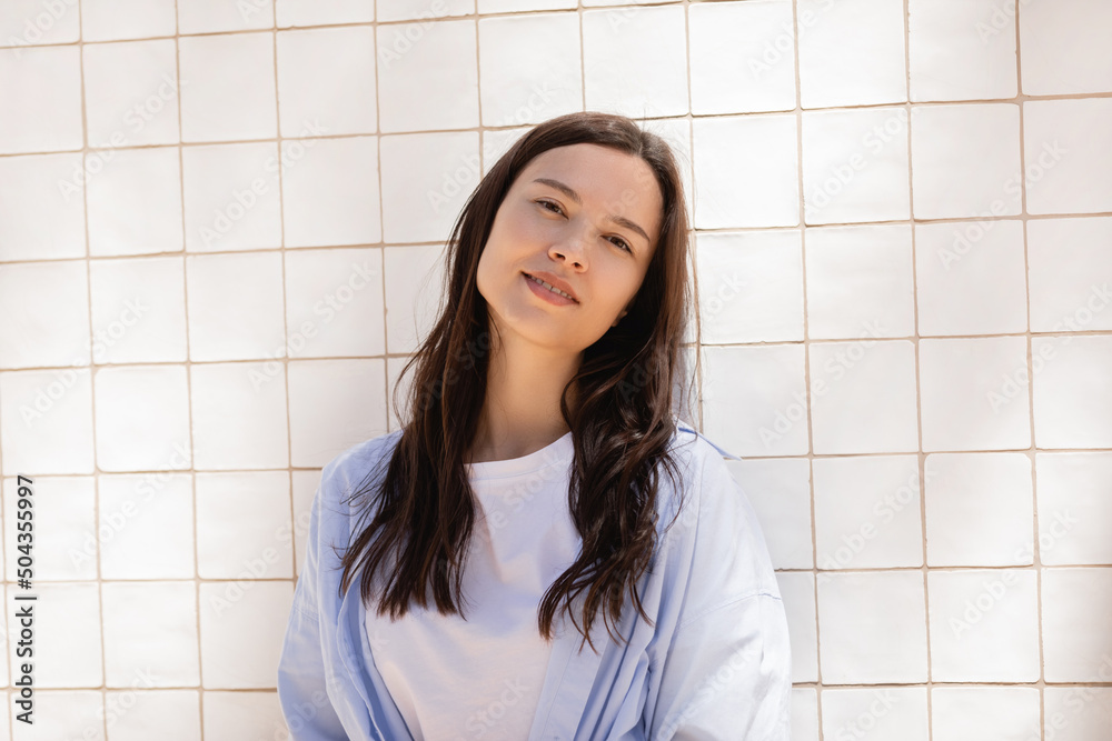 smiling brunette woman looking at camera while standing near white tiled wall. smiling brunette woman looking at camera while standing near white tiled wall.