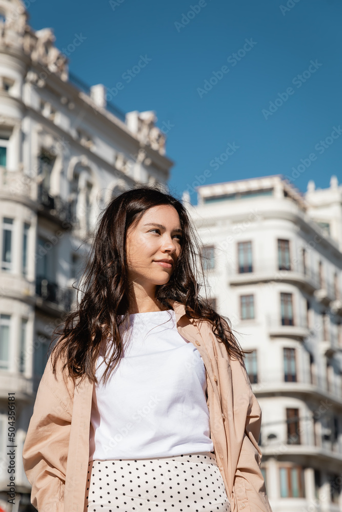 brunette woman smiling while standing on urban street and looking away.