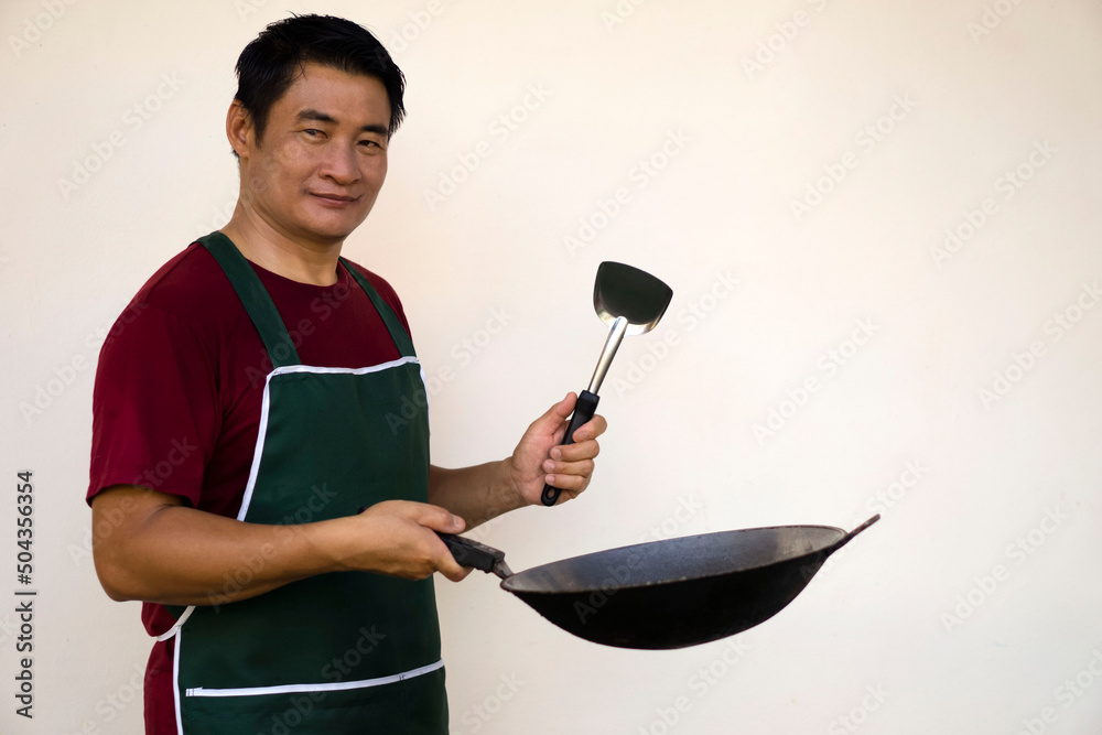 Portrait of handsome Asian man wears apron, holds cooking pan and ladle  spatula for frying. Look at camera, feels confident, isolated on white  background. Concept : love cooking. Kitchen lifestyle. Stock Photo