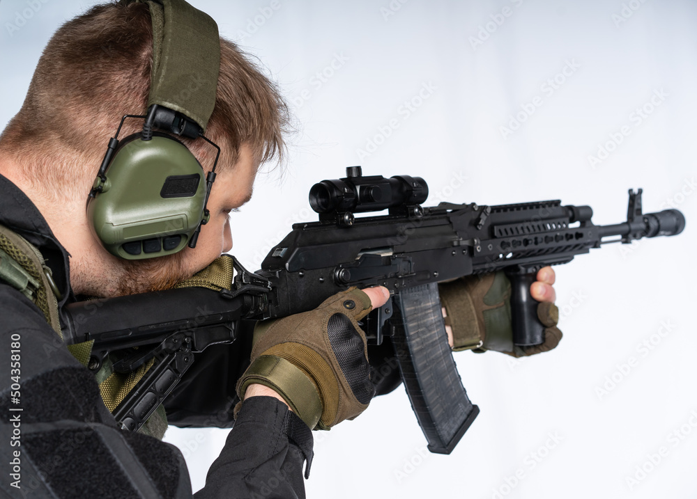 an airsoft soldier in full gear with his back to us is aiming at an optical sight. a man in an outfit, in headphones, a bulletproof vest, with a backpack and a belt. White background. an airsoft soldier in full gear with his back to us is aiming at an optical sight. a man in an outfit, in headphones, a bulletproof vest, with a backpack and a belt. White background.