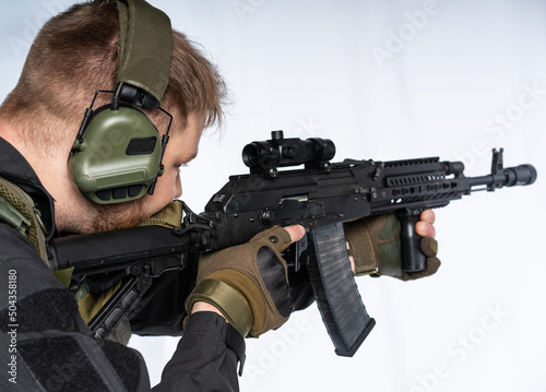 an airsoft soldier in full gear with his back to us is aiming at an optical sight. a man in an outfit, in headphones, a bulletproof vest, with a backpack and a belt. White background.