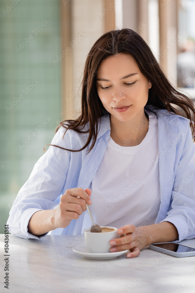 brunette woman holding teaspoon near coffee cup and smartphone with blank screen on cafe terrace. brunette woman holding teaspoon near coffee cup and smartphone with blank screen on cafe terrace.