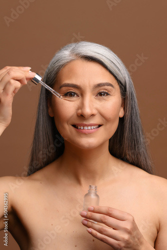 Vertical portrait of smiling middle-aged Asian woman with long gray hair holding jar with serum in hands  using pipette for applying cosmetology product