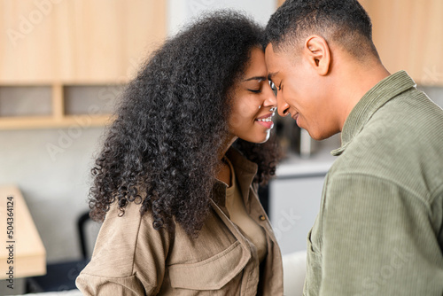 Lovely multiracial couple touching with foreheads in cozy modern kitchen at home. Smiling guy is bonding to his charming girl. Love and affection concept