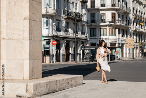full length of brunette woman in skirt walking along urban street.