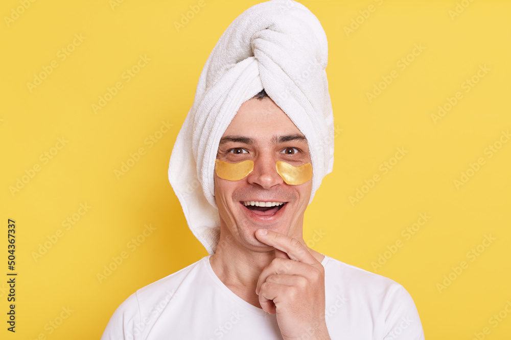 Smiling positive man wrapped towel on head doing morning cosmetology procedures, posing with patches under eyes isolated over yellow background, looking at camera with happy expression. Smiling positive man wrapped towel on head doing morning cosmetology procedures, posing with patches under eyes isolated over yellow background, looking at camera with happy expression.