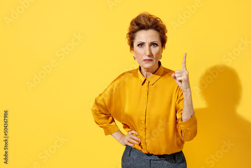 Elderly strict woman point index finger up, warns, notifies, recommends something. Photo of attractive woman in yellow shirt on yellow background photo