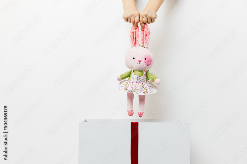 beautiful children's hands put a toy pink bunny in a white box with a red ribbon on an isolated white background. concept of gift, surprise
