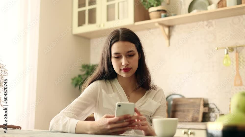 Close up attractive caucasian woman hold smartphone, watching social media sitting at the kitchen. Smiling young woman texting on her phone. Close up attractive caucasian woman hold smartphone, watching social media sitting at the kitchen. Smiling young woman texting on her phone.