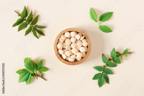 Directly Above Shot Of Pills and green leaves. Tablets in wooden bowl on beige neutral colors background. Homeopathic medicine. Organic medical capsules with herbal plant. Table top view. Copy space © Marina Demidiuk