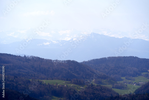 Panoramic view from local mountain Uetliberg with valley, village, agricultural fields and Swiss Alps in the background on a blue cloudy spring day. Photo taken April 14th, 2022, Zurich, Switzerland.