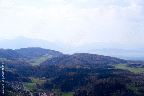 Fototapeta Naklejka Na Ścianę i Meble -  Panoramic view from local mountain Uetliberg with valley, village, agricultural fields and Swiss Alps in the background on a blue cloudy spring day. Photo taken April 14th, 2022, Zurich, Switzerland.
