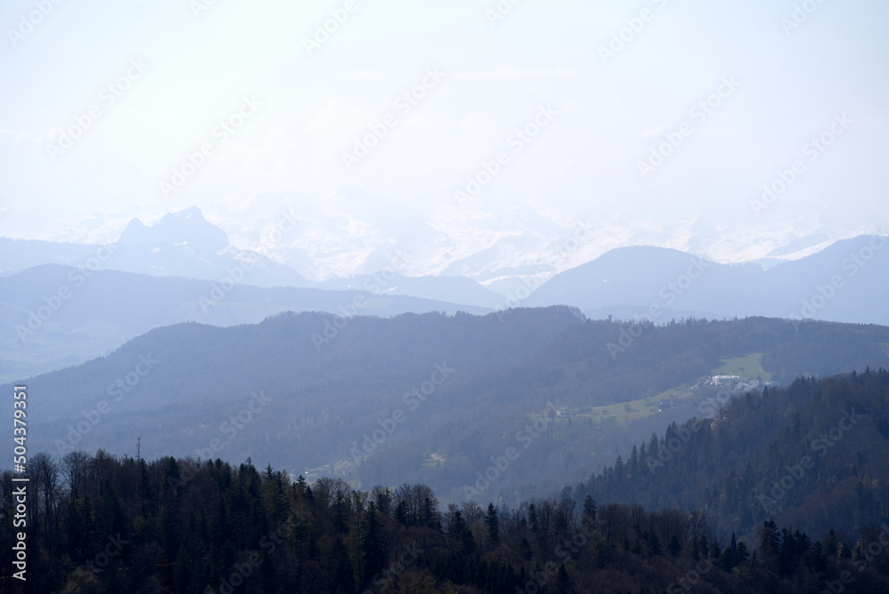 Obraz premium Panoramic view from local mountain Uetliberg with valley, village, agricultural fields and Swiss Alps in the background on a blue cloudy spring day. Photo taken April 14th, 2022, Zurich, Switzerland.