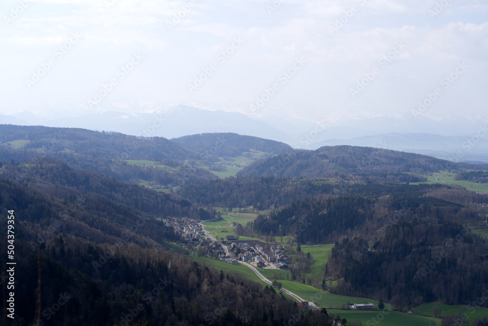 Panoramic view from local mountain Uetliberg with valley, village, agricultural fields and Swiss Alps in the background on a blue cloudy spring day. Photo taken April 14th, 2022, Zurich, Switzerland.