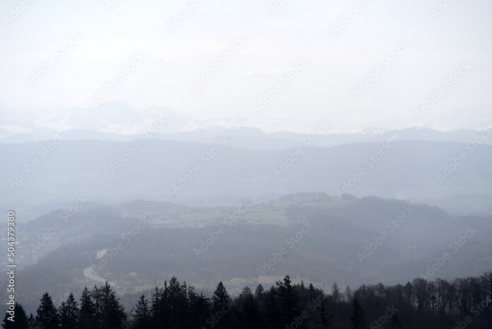 Obraz premium Panoramic view from local mountain Uetliberg with valley, village, agricultural fields and Swiss Alps in the background on a blue cloudy spring day. Photo taken April 14th, 2022, Zurich, Switzerland.