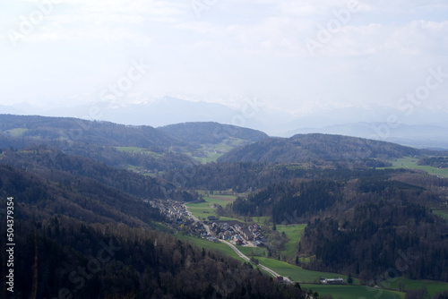 Panoramic view from local mountain Uetliberg with valley, village, agricultural fields and Swiss Alps in the background on a blue cloudy spring day. Photo taken April 14th, 2022, Zurich, Switzerland.