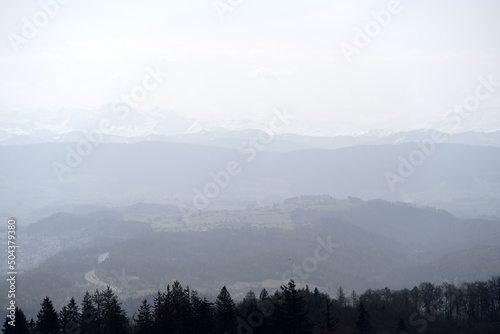 Panoramic view from local mountain Uetliberg with valley  village  agricultural fields and Swiss Alps in the background on a blue cloudy spring day. Photo taken April 14th  2022  Zurich  Switzerland.