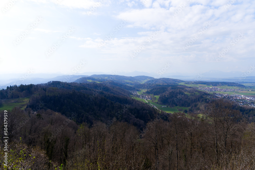 Panoramic view from local mountain Uetliberg with valley, village, agricultural fields and Swiss Alps in the background on a blue cloudy spring day. Photo taken April 14th, 2022, Zurich, Switzerland.