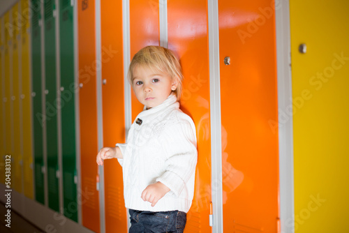 Sweet blonde toddler boy standing in front of a lockers in kindergarden or school hallway