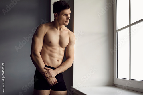 Portrait of handsome young man with sporty body posing in underwear standing by window. Caucasian man in comfortable black cotton underpants standing with pensive facial expression looking out window.