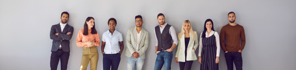 Young diverse fashion models posing against gray background. Banner with group portrait of confident young multi ethnic people in smart casual office wear standing in studio and leaning on grey wall
