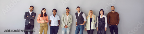 Young diverse fashion models posing against gray background. Banner with group portrait of confident young multi ethnic people in smart casual office wear standing in studio and leaning on grey wall