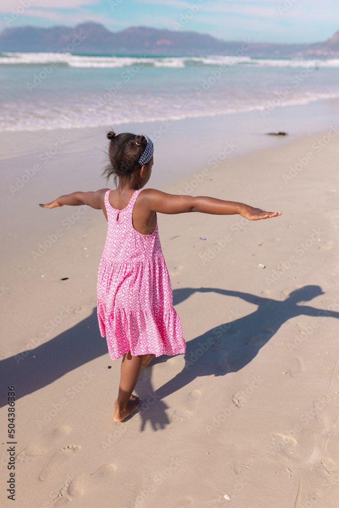 Rear view of playful african american girl with arms outstretched walking at beach against sky Rear view of playful african american girl with arms outstretched walking at beach against sky