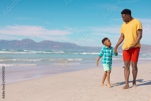 African american boy holding young father's hands while walking on sand at beach against blue sky