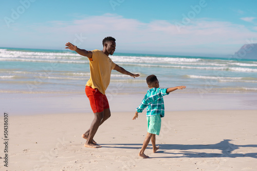 Playful african american young father and son with arms outstretched walking at beach against sky